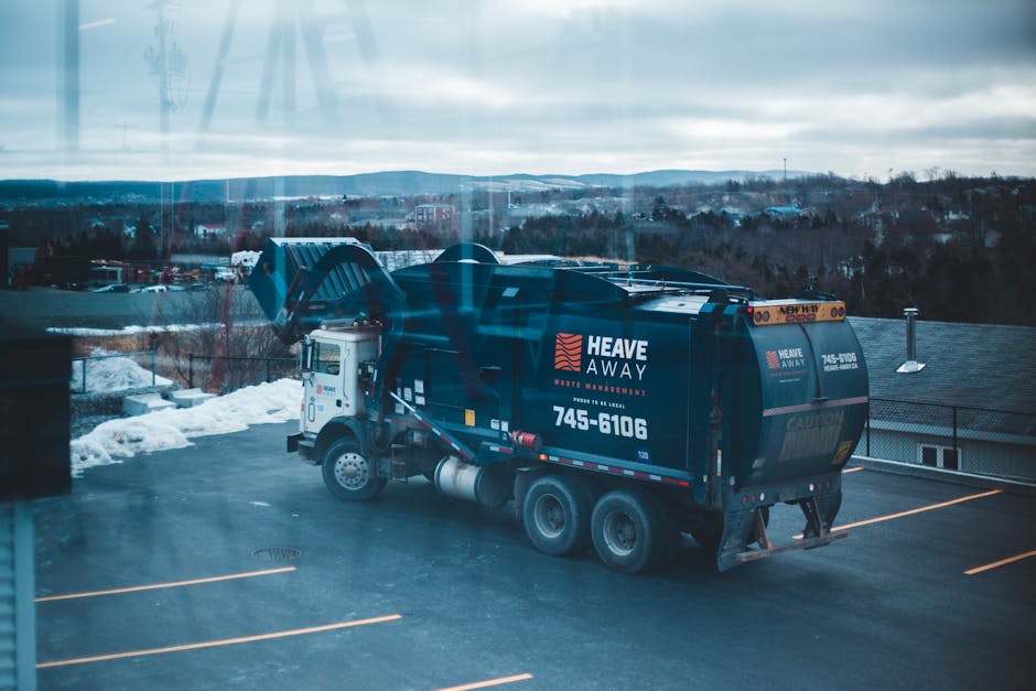 A large black waste disposal truck with the branding 'Heave Away' and contact number 745-6106 is parked in an outdoor lot during daytime, with snow on the ground and a cloudy sky overhead. The truck is positioned near a building, and the rear hatch is closed. In the background, there are distant hills and some industrial or commercial structures, indicating a semi-rural or suburban environment. The scene captures the truck ready for waste collection or disposal as part of a home relocation or decluttering service, with no visible people or loading activities occurring at this moment. The image is associated with professional removals and waste management services provided by Man with Van Penge, supporting efficient moving and disposal processes.