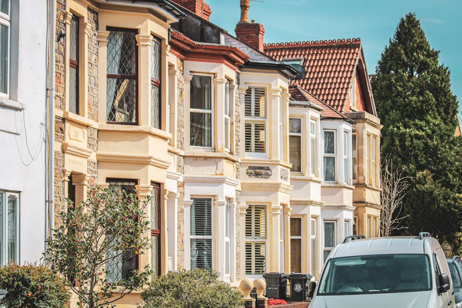 A row of traditional terrace houses with brick and cream-colored stone facades, large bay windows with decorative glass panes, and red-tiled roofs, situated on a residential street. In the foreground, a white van belonging to Man with Van Penge is parked on the pavement, positioned for home relocation or furniture transport, with its front facing the camera. The van is adjacent to an open doorway, where a removal team has begun loading a variety of packing boxes wrapped in plastic film and cardboard. The boxes vary in size and are being carried on a trolley toward the vehicle, with some already stacked inside or close to the van. The scene is illuminated by natural daylight, highlighting the details of the historic architecture and the moving process, which reflects house removals and packing activities common in professional relocation services referenced by Bromley Council permits for removals.