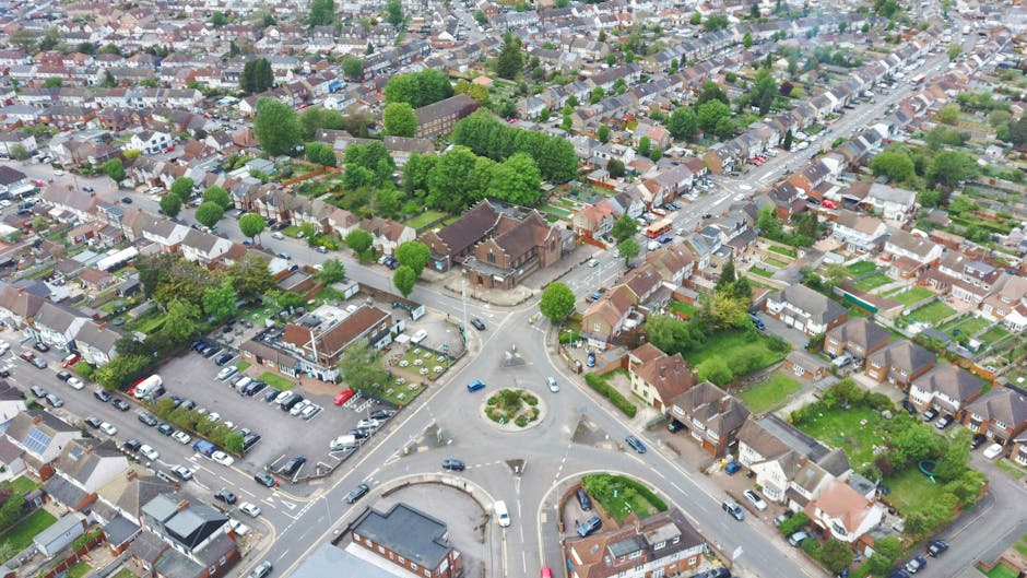An aerial view of a residential neighbourhood in Penge featuring a roundabout at the intersection of several streets. Surrounding the roundabout are small to mid-sized houses with gardens, some with visible fences and greenery. Parked cars occupy multiple parking areas and street spaces adjacent to the houses. Near the centre, there is a commercial building with parking spaces filled with vehicles. The scene includes trees lining the streets and within the neighbourhood, creating a lush environment. The lighting indicates daytime with natural sunlight illuminating the area, highlighting the details of the roads, vehicles, and residential properties. This image illustrates the typical layout of a suburban area suitable for home relocation or furniture transport. A professional removal service like Man with Van Penge might utilise such areas for loading or unloading household possessions during a move, emphasizing the importance of permits and planning in residential moving processes.