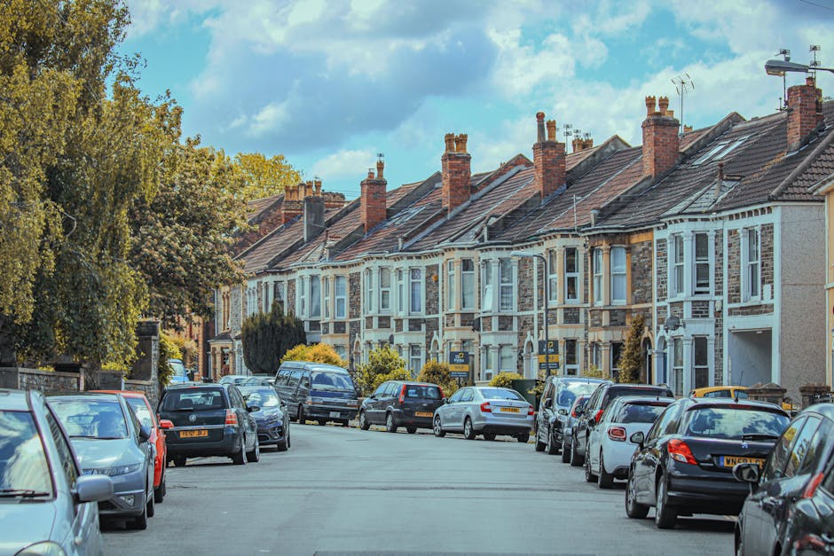 A row of traditional terrace houses with brick and cream-colored stone facades, large bay windows with decorative glass panes, and red-tiled roofs, situated on a residential street. In the foreground, a white van belonging to Man with Van Penge is parked on the pavement, positioned for home relocation or furniture transport, with its front facing the camera. The van is adjacent to an open doorway, where a removal team has begun loading a variety of packing boxes wrapped in plastic film and cardboard. The boxes vary in size and are being carried on a trolley toward the vehicle, with some already stacked inside or close to the van. The scene is illuminated by natural daylight, highlighting the details of the historic architecture and the moving process, which reflects house removals and packing activities common in professional relocation services referenced by Bromley Council permits for removals.