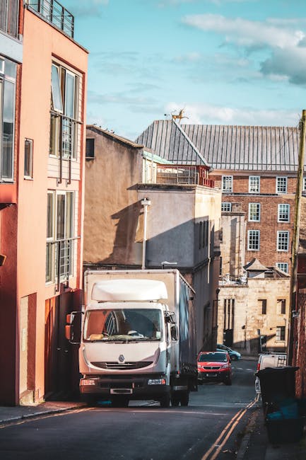 A white moving van parked on a narrow residential street with multi-storey brick and stucco buildings on either side. The van is positioned close to the sidewalk, with its front facing slightly to the