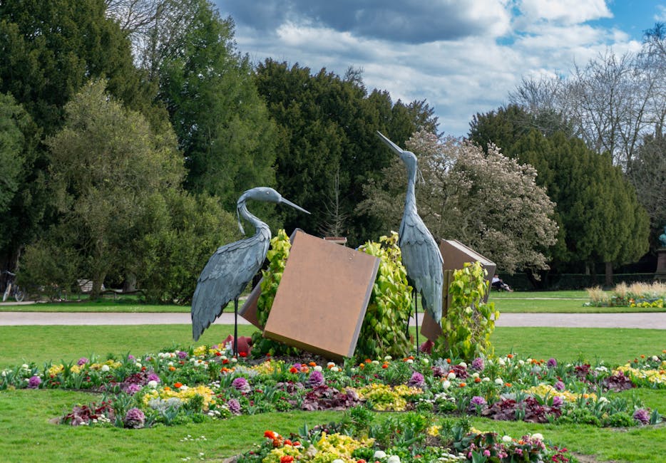 A flower bed featuring vibrant, blooming flowers surrounds two metal crane sculptures with long necks and pointed beaks, positioned in a park setting with lush green trees, some with white blossoms, in the background; the sculptures are partially obscured by cardboard packing and green foliage, indicating an ongoing home relocation process involving packing and transport of outdoor decorative items, as seen near a paved walkway under natural daylight, with a clear sky overhead; the scene is part of a house removal and furniture transport operation executed by Man with Van Penge, who handles removals and moving logistics across residential areas.