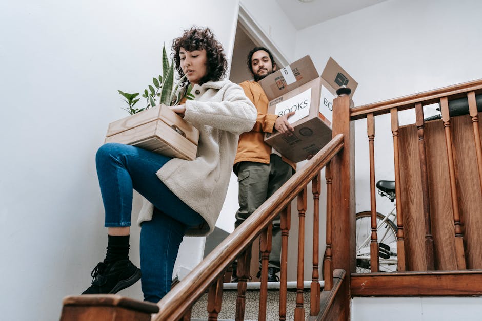 A woman with curly hair, dressed in a cream coat, blue jeans, and black shoes, is lifting a cardboard box with a potted plant on top up a staircase inside a home. Behind her, a man with dark hair and a beard, wearing a brown jacket, is carrying several stacked cardboard boxes labeled with 'BOOKS' and other items, preparing for a home relocation. The staircase features wooden handrails and spindles, with a bicycle partially visible on the landing. The environment is well-lit, and the scene depicts a typical moving process with packing supplies and furniture being transported. This visual aligns with furniture transport and packing during house removals, as handled by Man with Van Penge, reflecting the logistical aspects of staircase rules for flat moves.