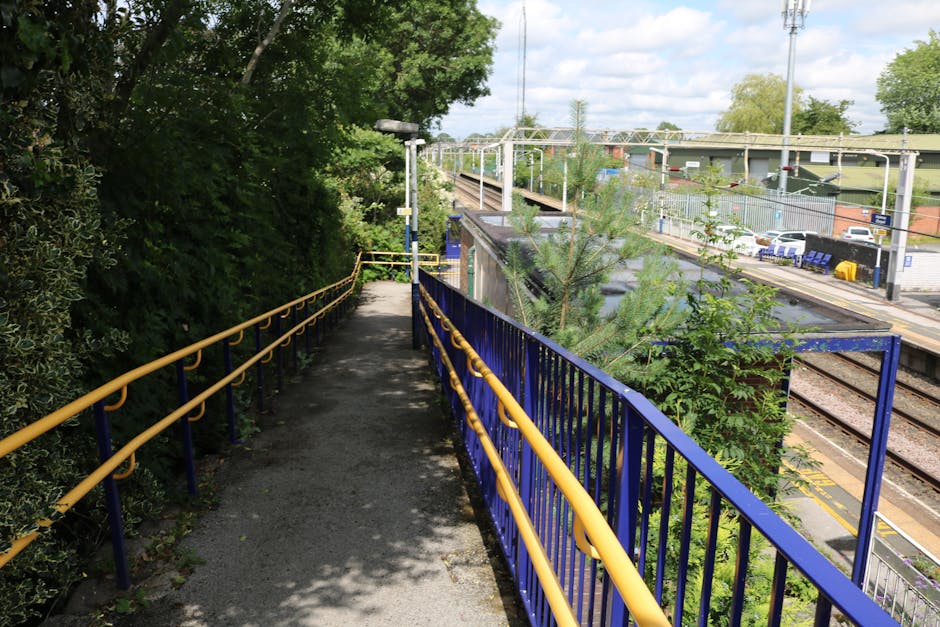 A narrow, outdoor footpath with a metal railing painted in blue and yellow, running alongside a railway station platform. The path is shaded by lush green trees and bushes on the left side, while on the right side, the station tracks and platform are visible, with parked cars, benches, and station signage in the background. The ground is paved with asphalt, and a tall lamppost is positioned at the start of the path, which appears to be used for access during home relocation or furniture transport activities. The overall scene depicts the process of moving items from a property to a vehicle or storage area, with professional moving services like Man with Van Penge potentially utilising such pathways for efficient loading and transport related to house removals, packing, and logistical planning near Penge East Station.