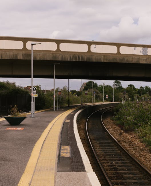 A quiet railway station platform with two parallel train tracks running through the scene, bordered by a concrete edge with tactile paving near the edge for accessibility. On the right side, there is a small brick building with a black roof, featuring a trash bin and a covered area for waiting or ticketing. The platform is mostly empty, with a few blue lamp posts evenly spaced along its length, and behind the station building, there are lush green trees and bushes, providing a natural backdrop. A footbridge with a white, perforated metal railing arches across the tracks, connecting the two sides of the station. The overall scene suggests a calm environment suitable for home relocation or furniture transport services, with visible elements that support the logistics of loading and unloading, such as the flat platform and open space. Man with Van Penge, a specialist in removals, might use this station for moving household items or furniture as part of an efficient packing and moving process in the Penge area.
