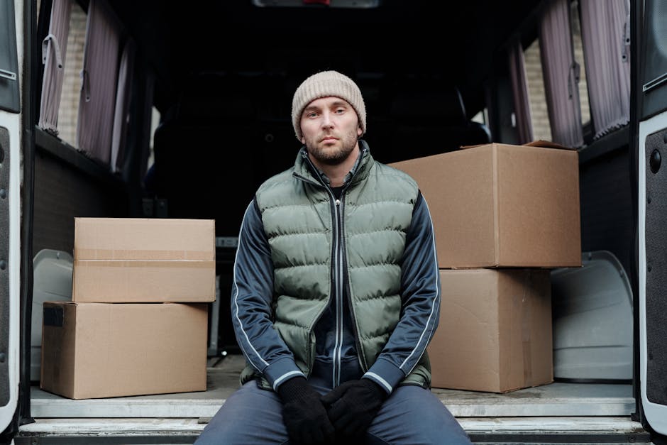 A man wearing casual clothing and a black cap is engaged in the loading process of house relocation, carrying three medium-sized cardboard boxes stacked in his arms towards a white commercial van park