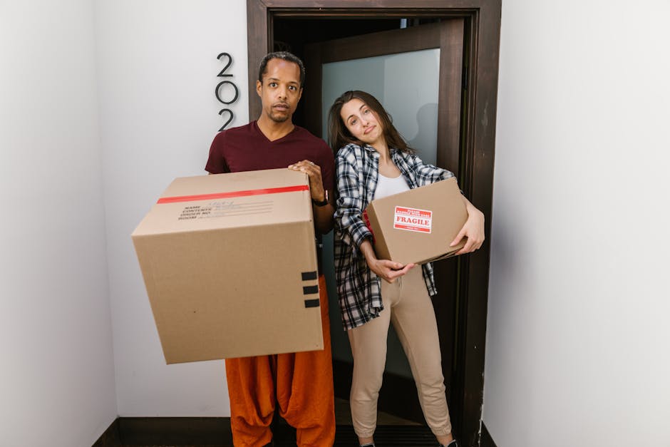 A man and a woman standing inside a doorway of a residential building, both holding cardboard boxes during a home relocation process. The man, with dark hair and wearing an orange and maroon outfit, holds a large box with black tape, while the woman, with long brown hair and dressed in a plaid shirt and beige pants, carries a smaller box marked with a red 'Fragile' label. They are positioned on a narrow hallway with plain white walls, and behind them is a dark wooden door frame. The scene takes place during daylight, with natural light illuminating the area, and the background suggests an indoor setting with visible packaging materials and the beginning of a moving or packing activity, indicating a furniture transport and moving process often coordinated by professional removals services like Man with Van Penge.