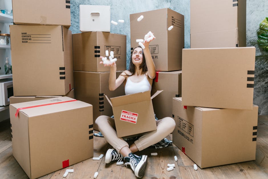 A young woman with long dark hair, wearing a white sleeveless top, beige pants, and black-and-white sneakers, is sitting on a wooden floor surrounded by numerous cardboard packing boxes, some labeled with 'Fragile.' She is inside a room with textured light blue walls, with a doorway visible behind her, as she joyfully tosses white packing foam peanuts into the air. The boxes are stacked and scattered around her, with some open and others sealed with red or black tape. To the left, part of a white countertop with kitchen items are visible, and a green plant is in the corner. The scene captures the packing process during home relocation, illustrating proper handling of packed items before loading onto a van or transport vehicle, which aligns with the services offered by Man with Van Penge in their removals category. Lighting appears natural, illuminating the room clearly, emphasizing the organized yet active packing environment connected to moving and storage solutions, especially when facing storage shortages in SE20.